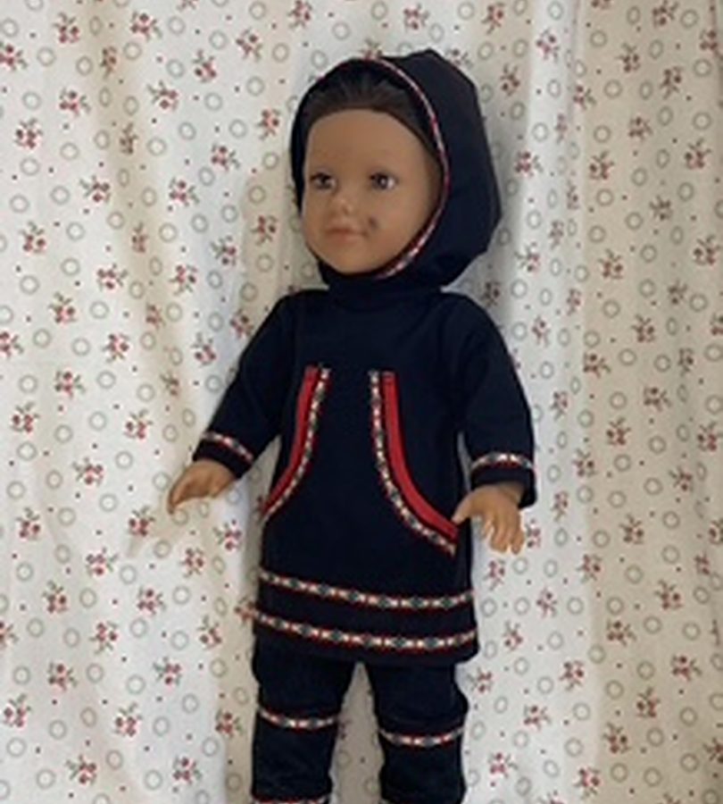 A doll dressed in a navy parka and trousers trimmed with ribbon.