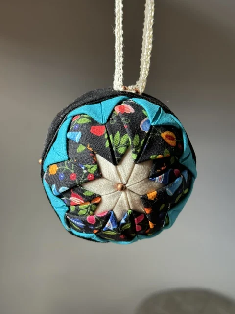 A teal and cream ornament with a braided cord hanger, laid out on a dark surface.
