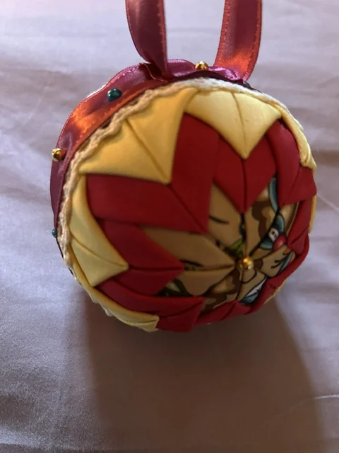 A red and gold ornament photographed close up on a white cloth, the ribbon hanger curving away to one side.