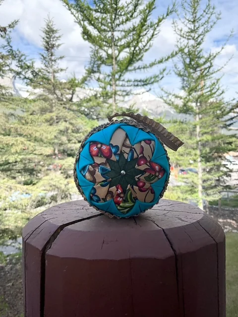A finished ornament set on top of a wooden post outdoors, with spruce trees and open sky behind it.