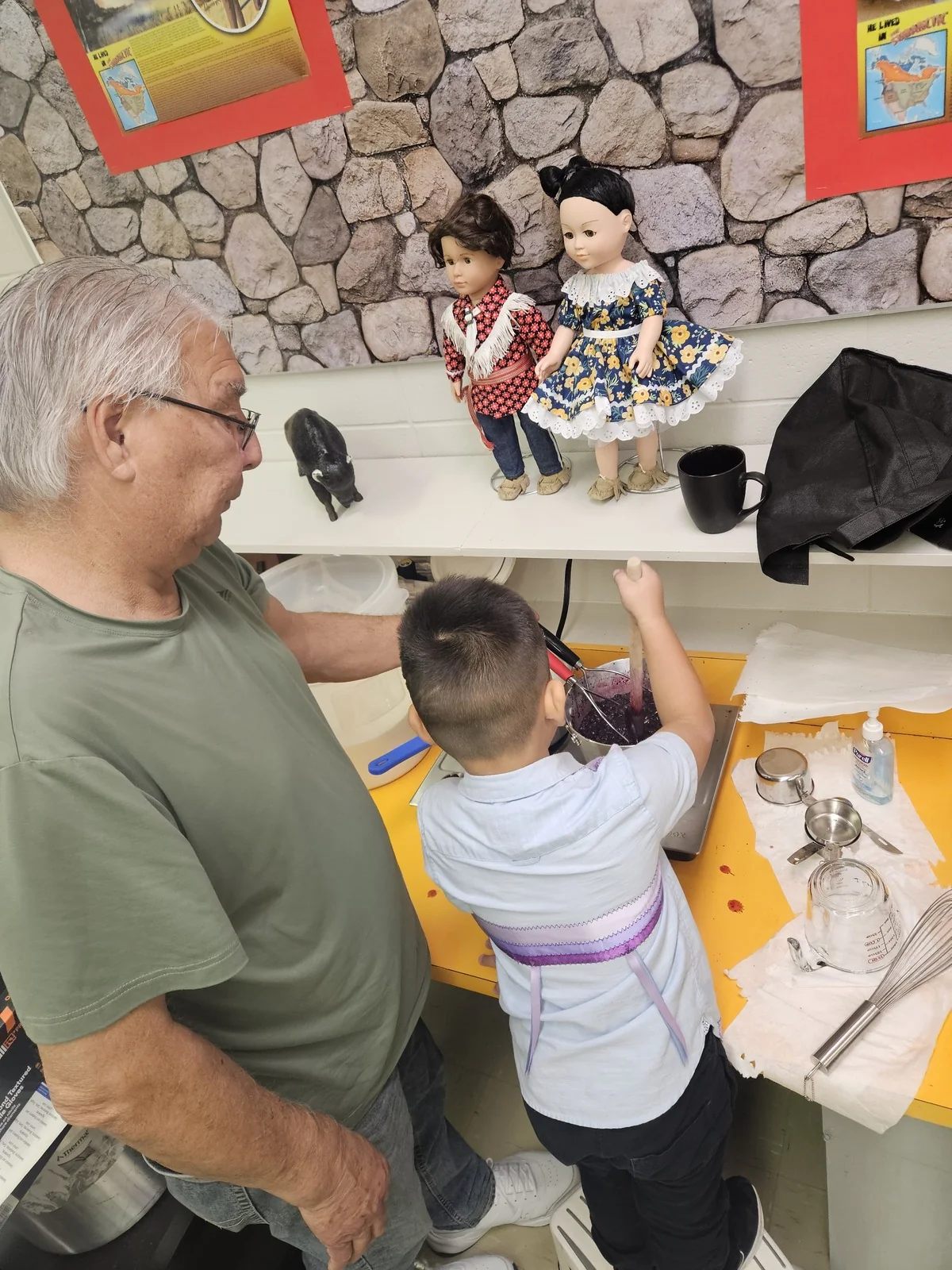 An Elder sitting with a child at a work table, dolls in regalia lined up on the ledge behind them.
