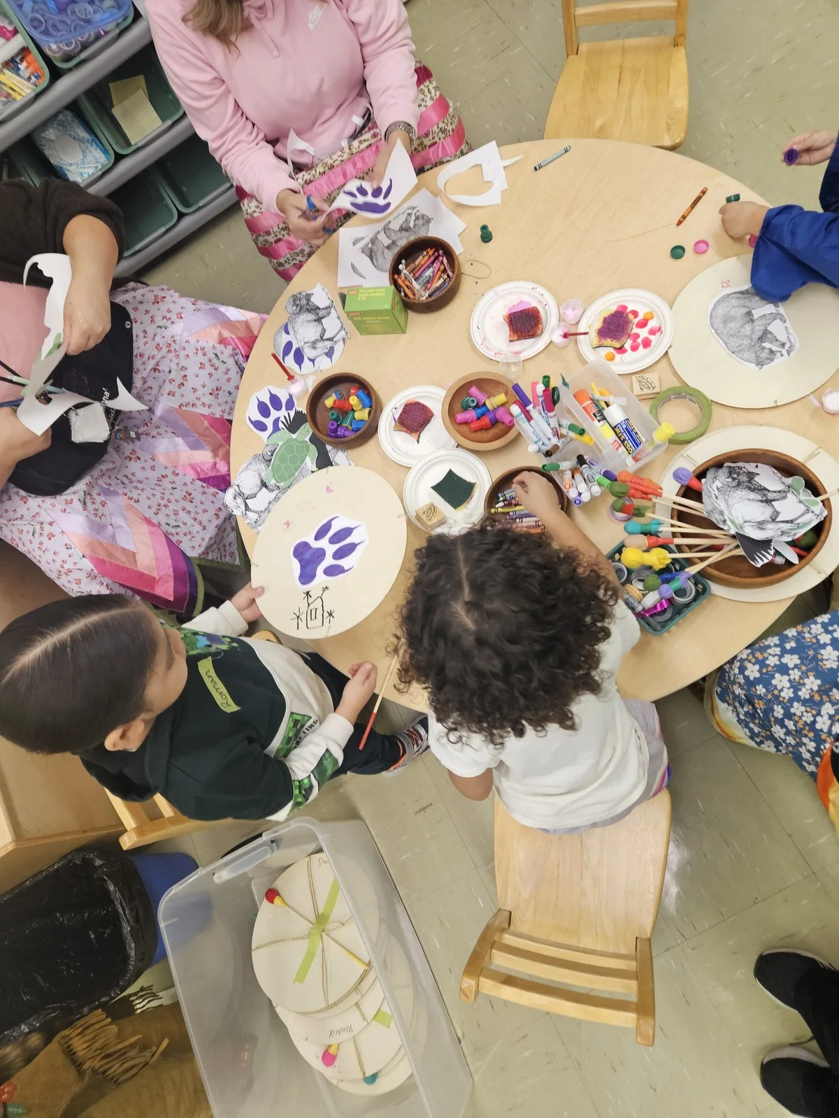 Children painting at a round table, seen from above, brushes and palettes spread between them.