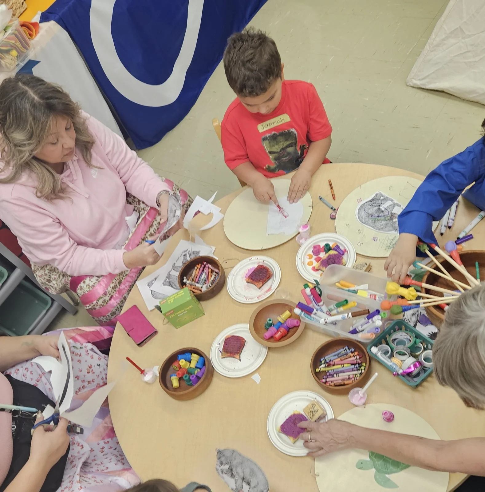 Children and an adult painting together at a table covered with paints and brushes.