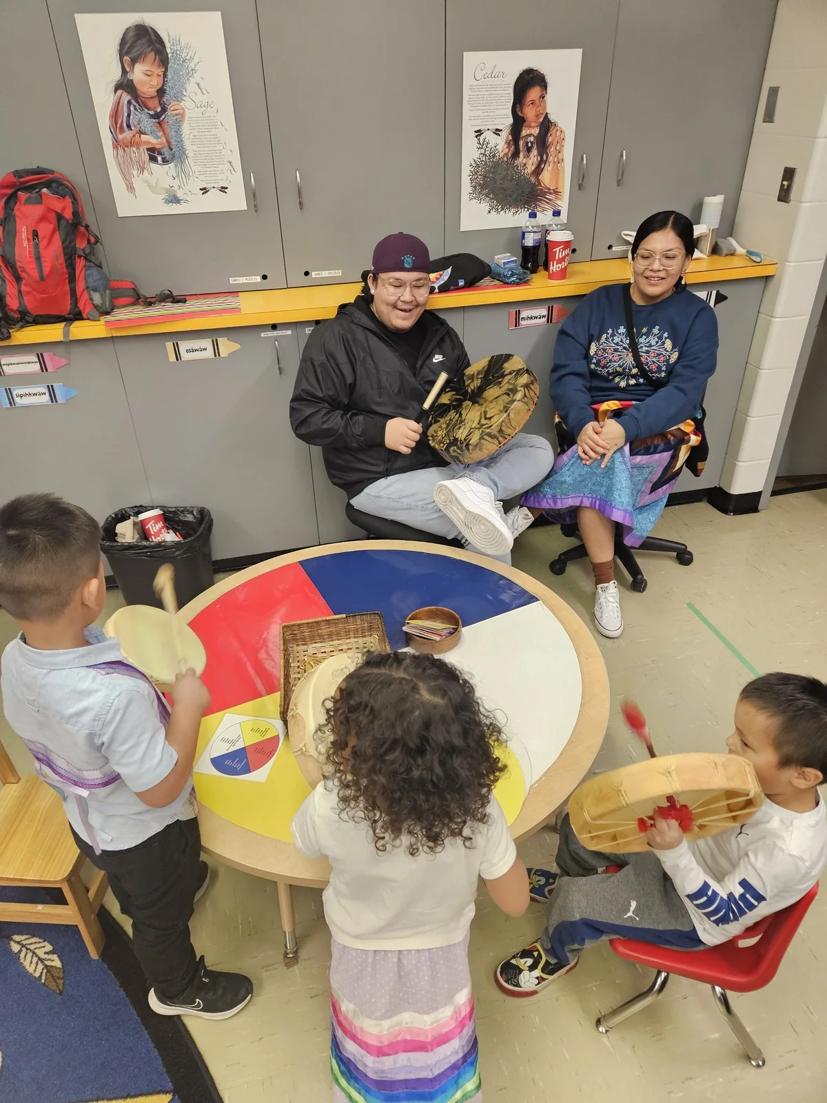 Two staff seated with hand drums while children gather at a low table in front of them.