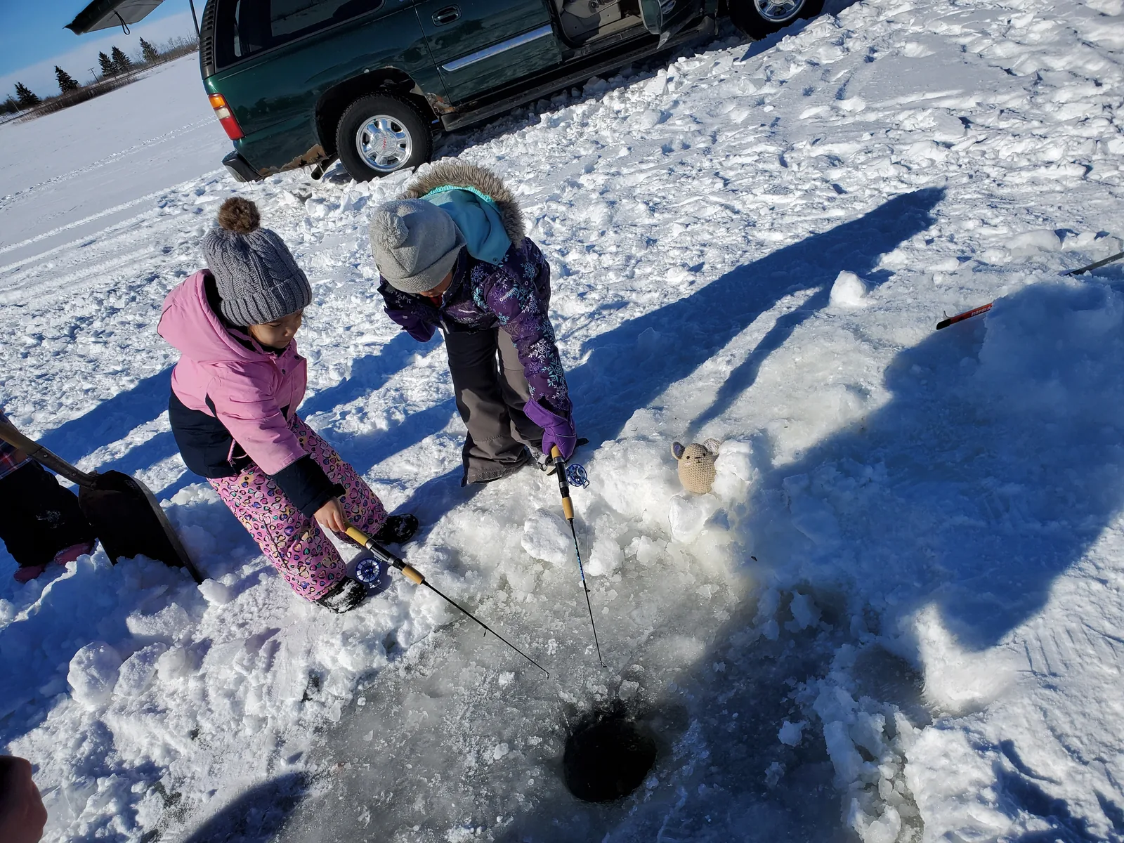 Two children in snowsuits leaning over a hole in the ice with a line in the water.