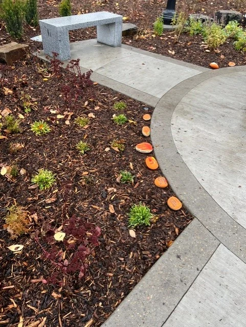 The painted stones set in a line along the edge of a planting bed beside a curved concrete path, a granite bench behind.