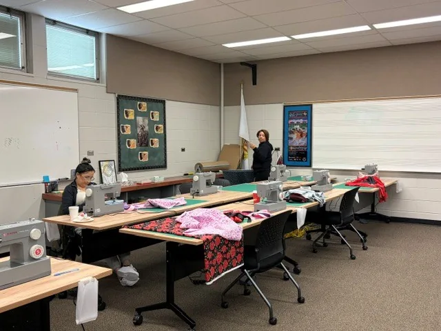 A sewing classroom set up for the class: rows of tables with sewing machines, green cutting mats and bundles of fabric, two people working at the far end.
