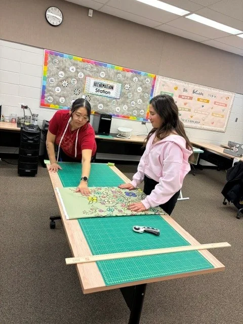 Two people smoothing a green floral skirt panel flat on the cutting mat, a rotary cutter and a yardstick on the table.