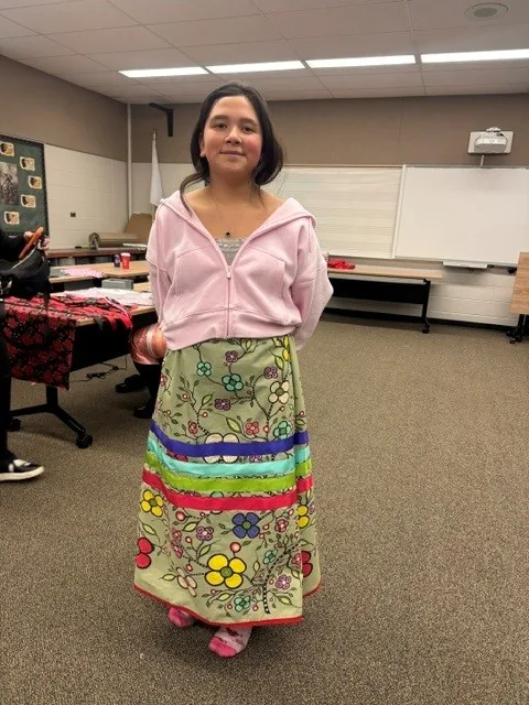 A young woman wearing her finished green floral ribbon skirt, standing in the middle of the classroom.