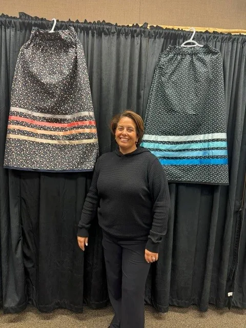 A woman standing between two finished skirts hung on a curtain behind her, one navy with pink bands, one dark with blue bands.